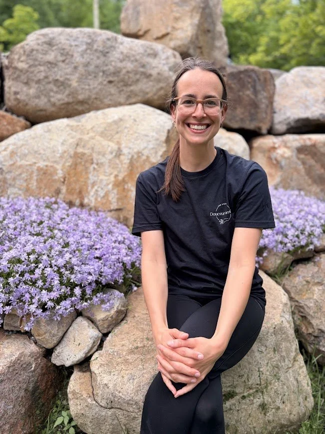 Femme souriante avec des lunettes, portant un t-shirt noir, assise sur des rochers avec des fleurs violettes et un paysage de nature en arrière-plan.