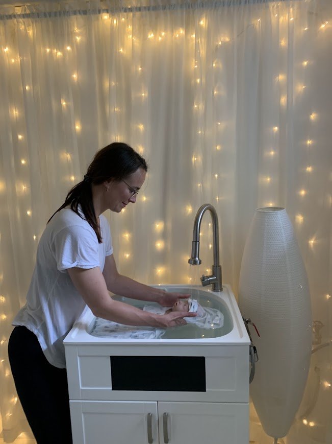 Une femme qui tient un bébé dans un bain pour bébé dans une pièce décorée avec des guirlandes lumineuses.