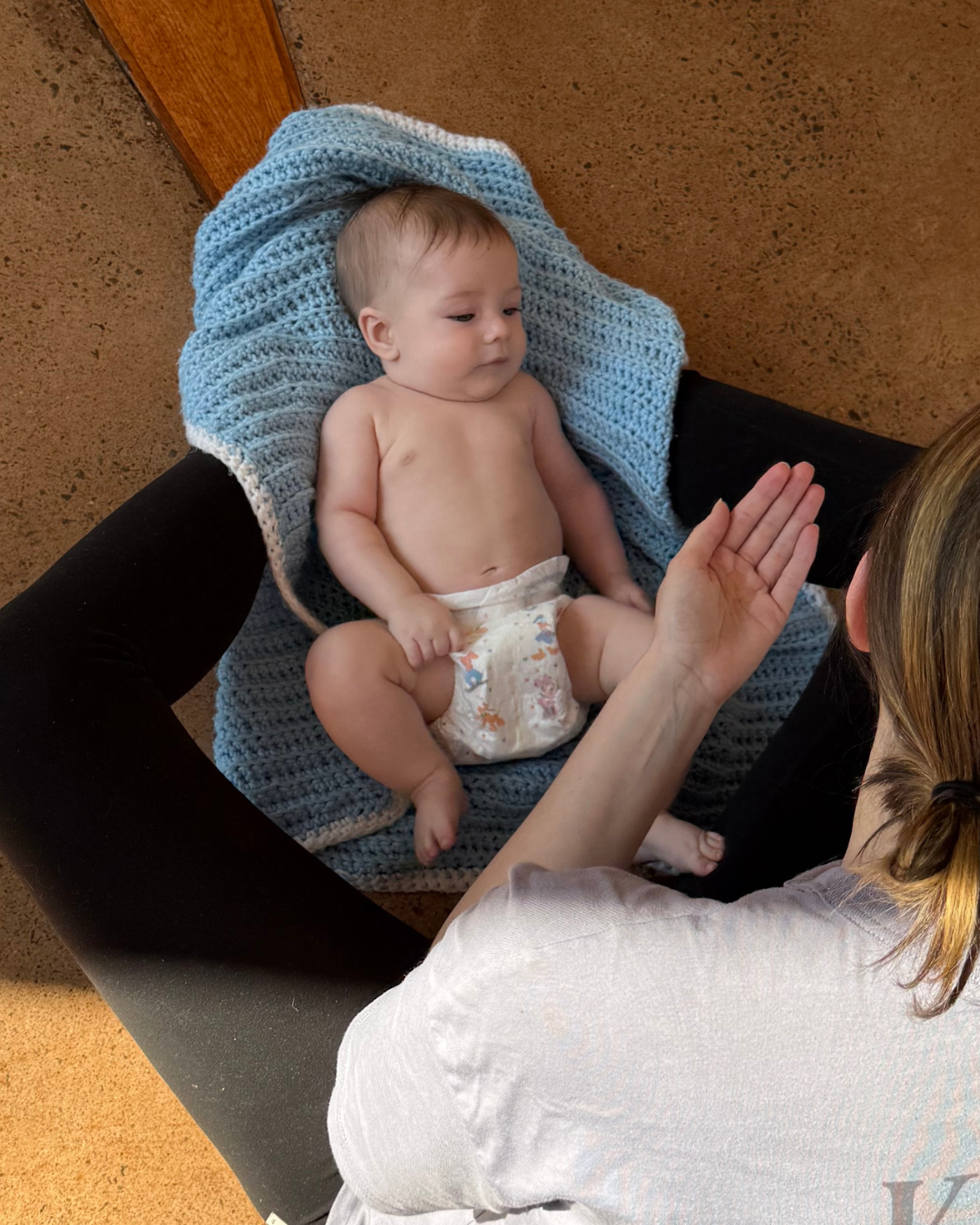 Un bébé allongé sur une couverture bleue, sans vêtements, en train de faire un geste de mains avec une femme dont la main est visible