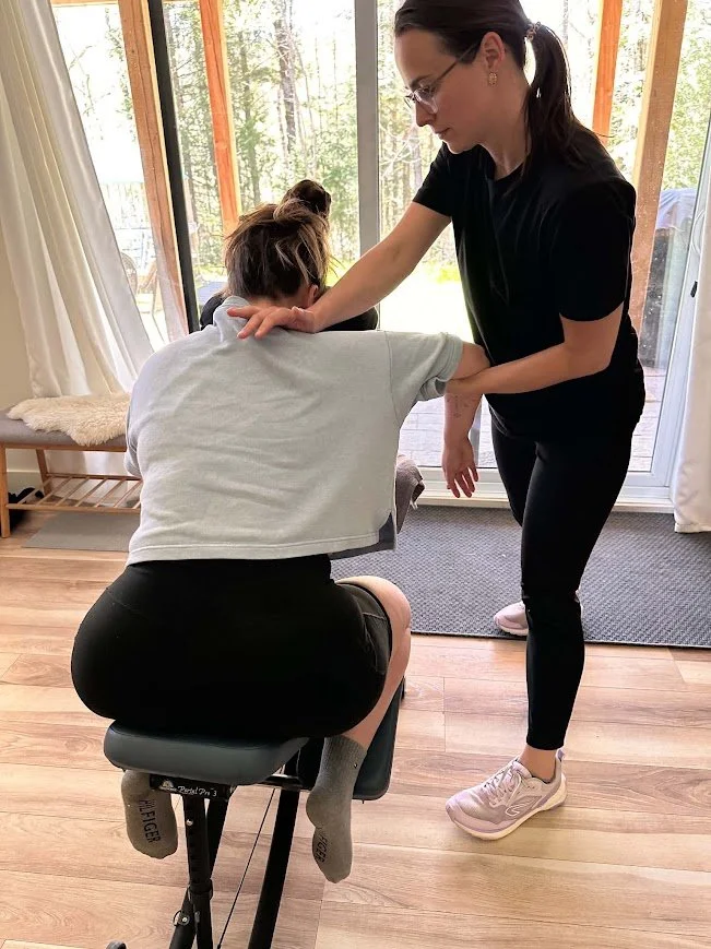 Une femme recevant un traitement ou une thérapie par un professionnel dans une pièce ensoleillée avec vue sur la nature. La femme est assise sur une table de massage, tandis que la thérapeute la soutient selon une position spécifique.