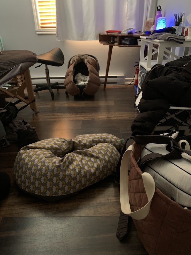 Une pièce avec un sol en bois, contenant un tapis pour animaux de compagnie, une maison pour chat, une table, une chaise, des étagères avec des objets, un chauffe-lit, et un sac. Il y a une fenêtre avec des volets en bois. La pièce semble être un espace de détente ou une zone pour animaux.