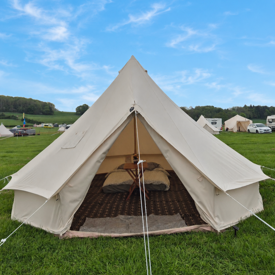 A large beige canvas tent set up on a grassy field, with a bed visible inside and a small wooden table. Other tents and cars are visible in the background of the festival, under a blue sky with scattered clouds.