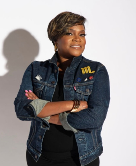 Woman with short hair wearing a denim jacket with embroidered patches, posing with arms crossed against a plain background.