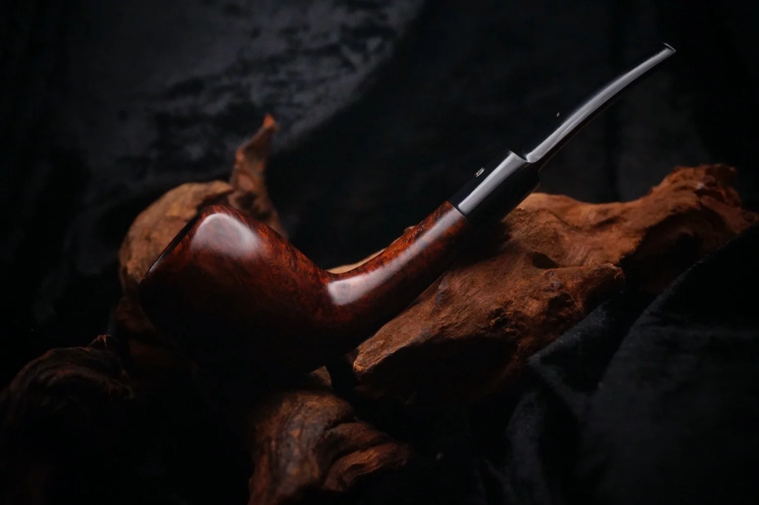 A smoking pipe with a curved wooden bowl and a black stem rests on a piece of driftwood against a dark background.