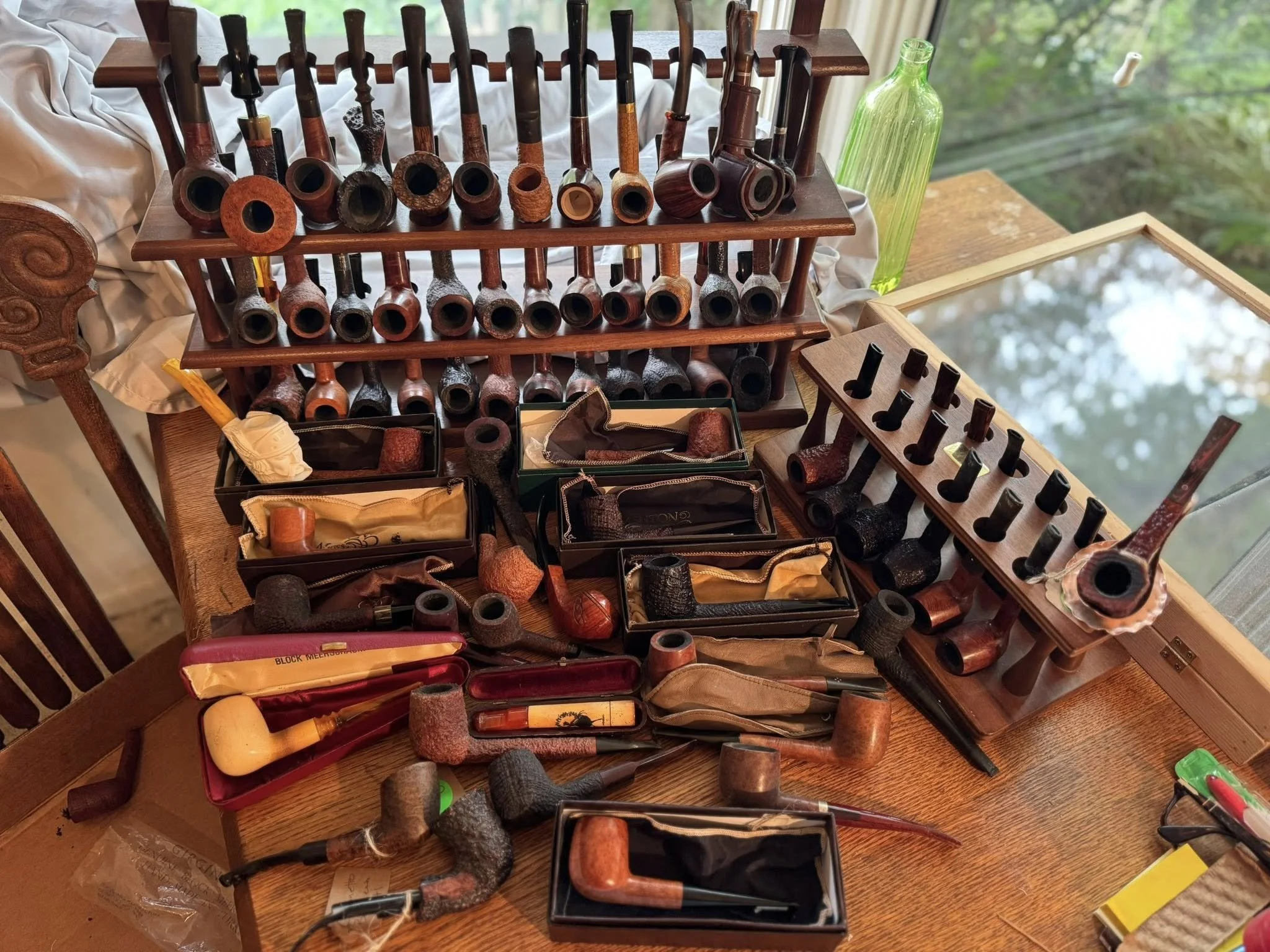 A display of various tobacco pipes arranged on a wooden table with some in open boxes and others on a wooden pipe rack, next to a green glass bottle and a mirror, with a window in the background showing greenery outside.
