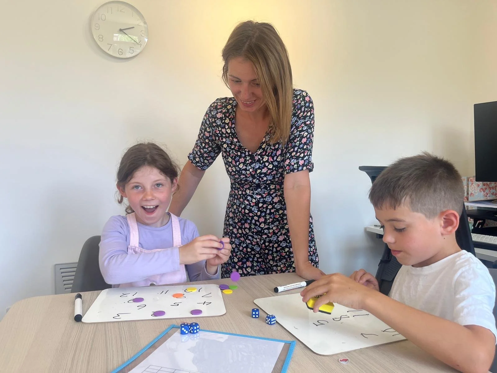 A teacher with two children playing a game at a table in a classroom. The girl on the left has brown hair, is smiling, and holds colorful game pieces. The boy on the right has short hair, looks focused, and handles game tokens. There are game boards, dice, and marker pens on the table. A clock on the wall shows 2:43.