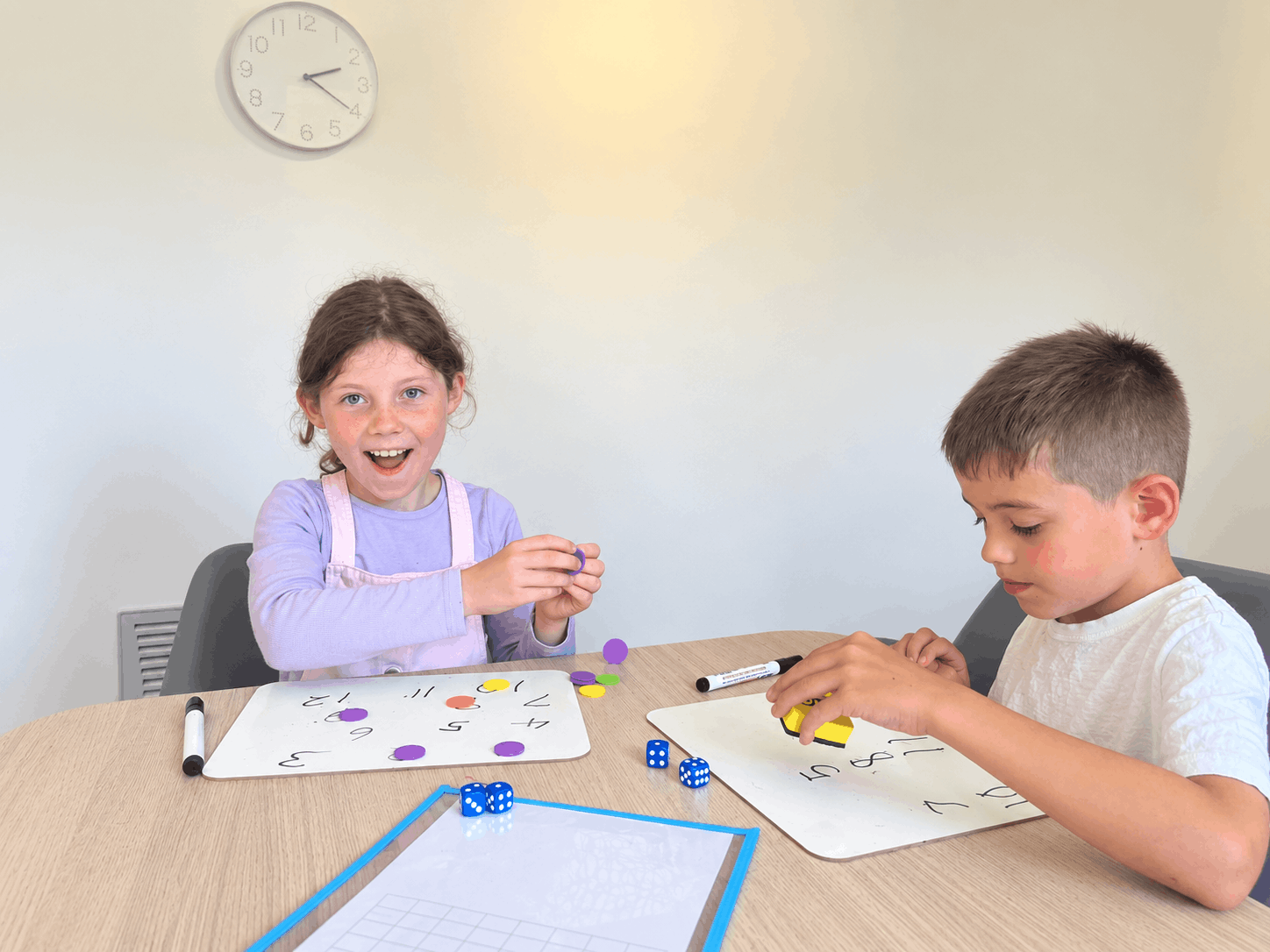 Two children playing a board game at a table in a room. The girl on the left has curly brown hair, a purple shirt, and is excited, while the boy on the right has short hair and is focused on the game. There are colorful game pieces and dice on the table.