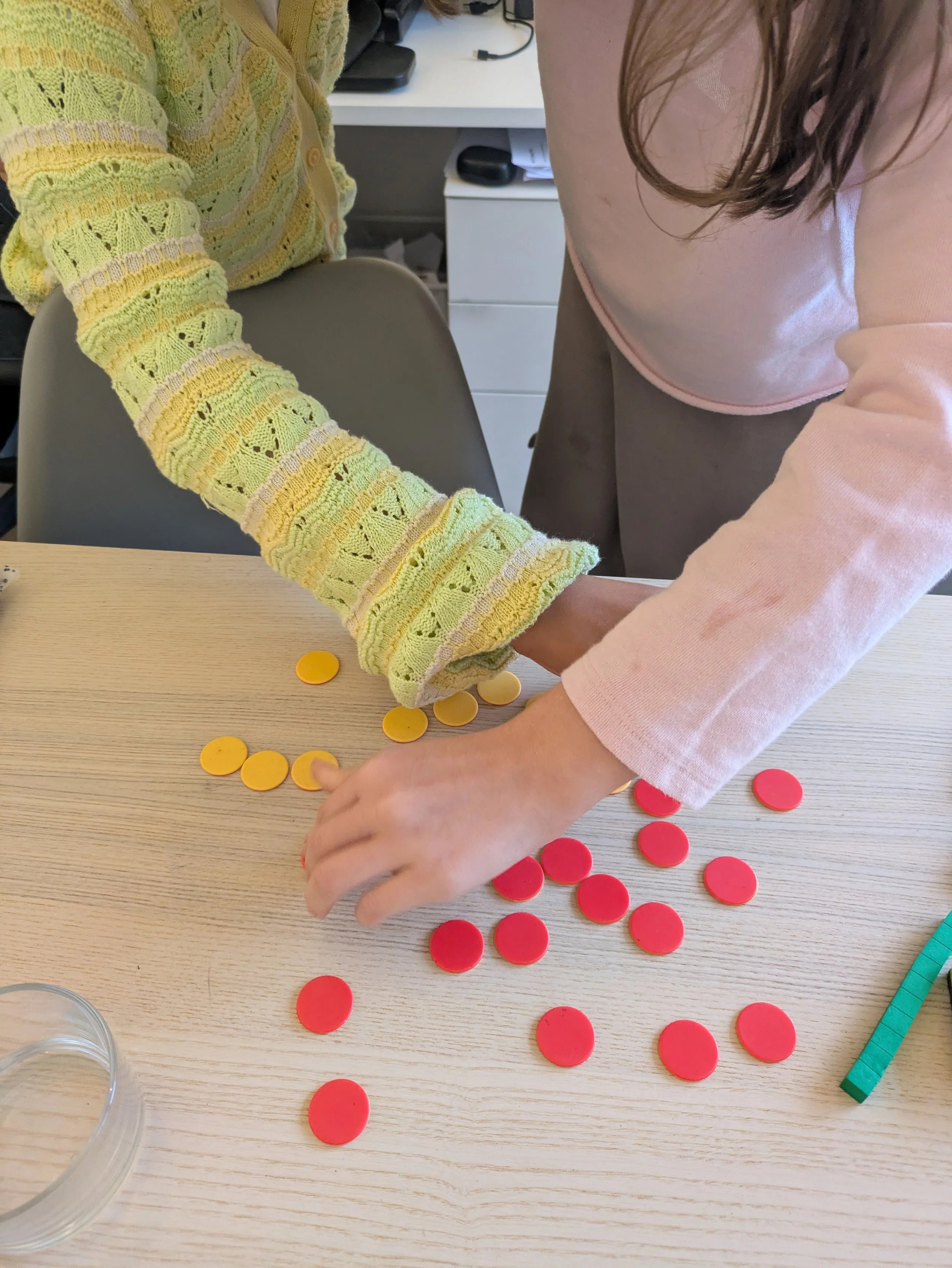 Two children are playing a game involving yellow and red circular tokens on a light-colored wooden table, with a measuring ruler visible on the right side.