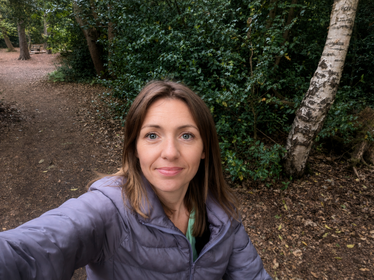 A woman taking a selfie outdoors on a forest trail with trees and greenery behind her.