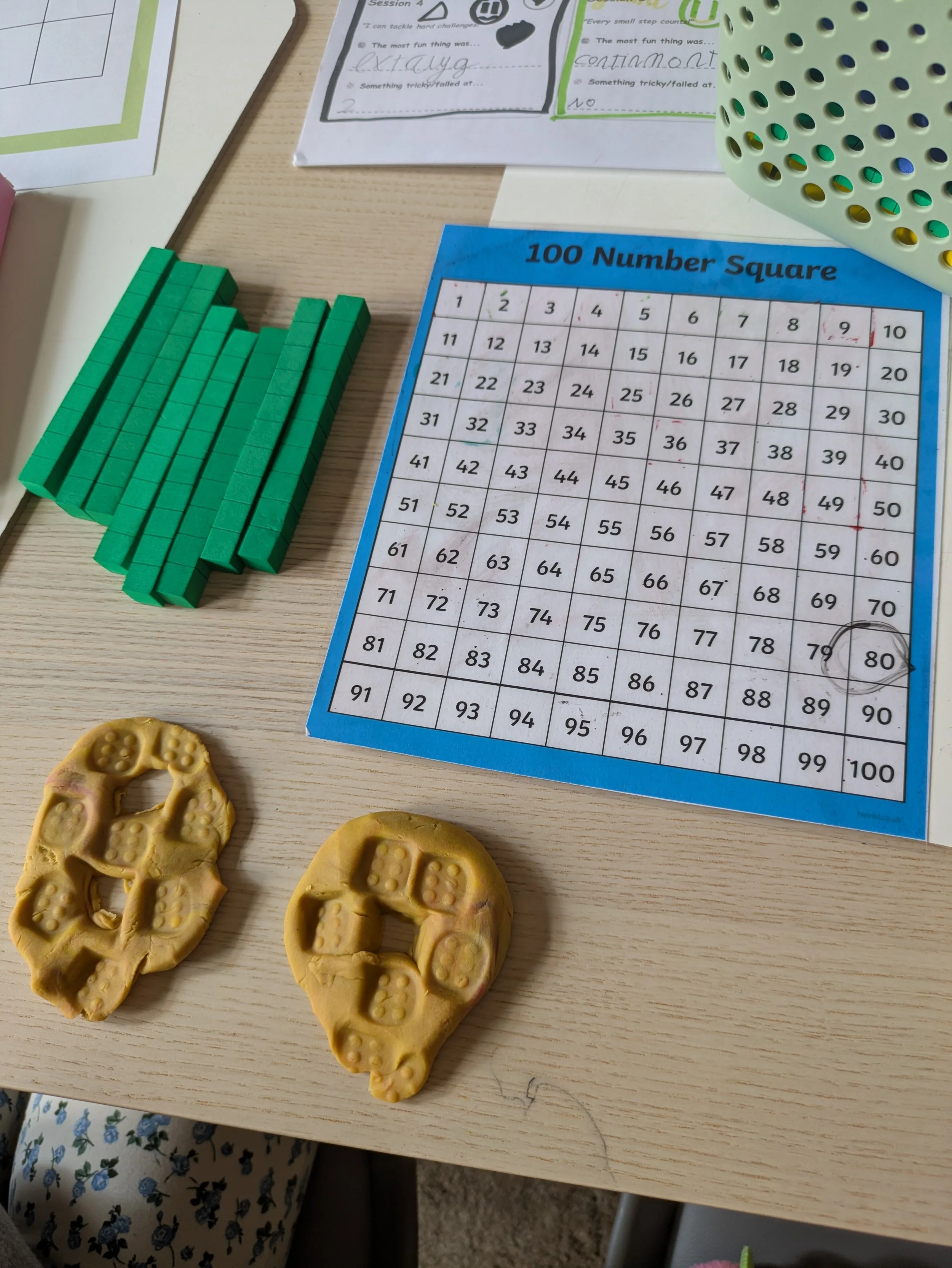 A wooden table holding a bingo card named '100 Number Square', green tiles arranged in a pattern, and beige cookies shaped like gloves with a printed pattern resembling dice faces.