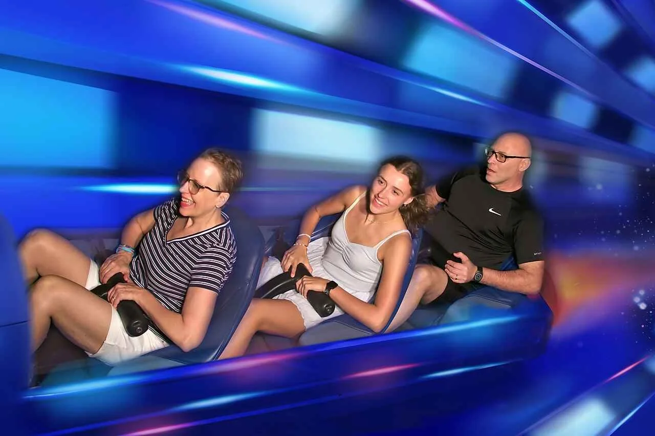 Three adults in a space mountain roller coaster car going through a tunnel of futuristic clue lights.