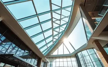 The image shows a modern interior with large glass windows and a geometric glass ceiling letting in natural sunlight.