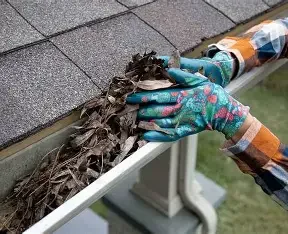 Person wearing colorful gloves cleaning leaves and debris from a gutter along a house roofline.