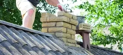 Person building a brick chimney on a house roof surrounded by greenery.