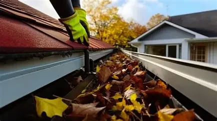 Person wearing gloves cleaning leaves out of a rain gutter on a house roof.