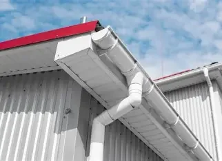 Close-up of an industrial white rain gutter and downspout attached to a building with metal siding, under a partly cloudy sky.