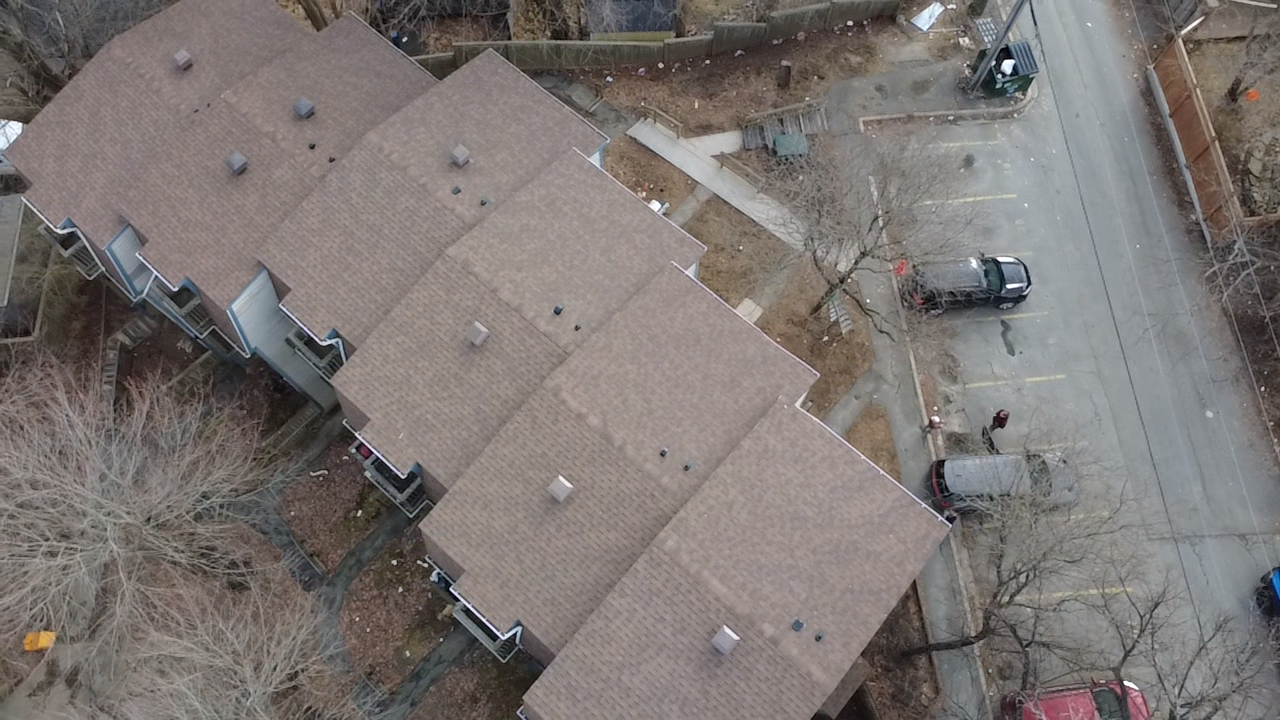 Aerial view of a multi-unit apartment complex with brown roofs, surrounded by trees and a parking lot with some parked cars.