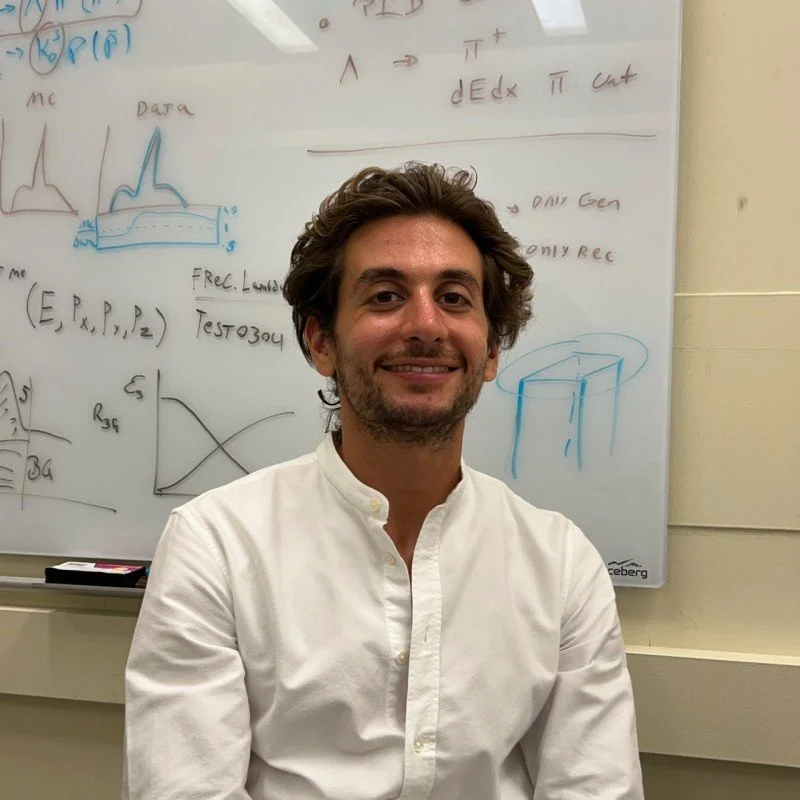 A smiling man with wavy brown hair, wearing a white button-up shirt, sitting in front of a whiteboard covered in handwritten notes, equations, and diagrams.