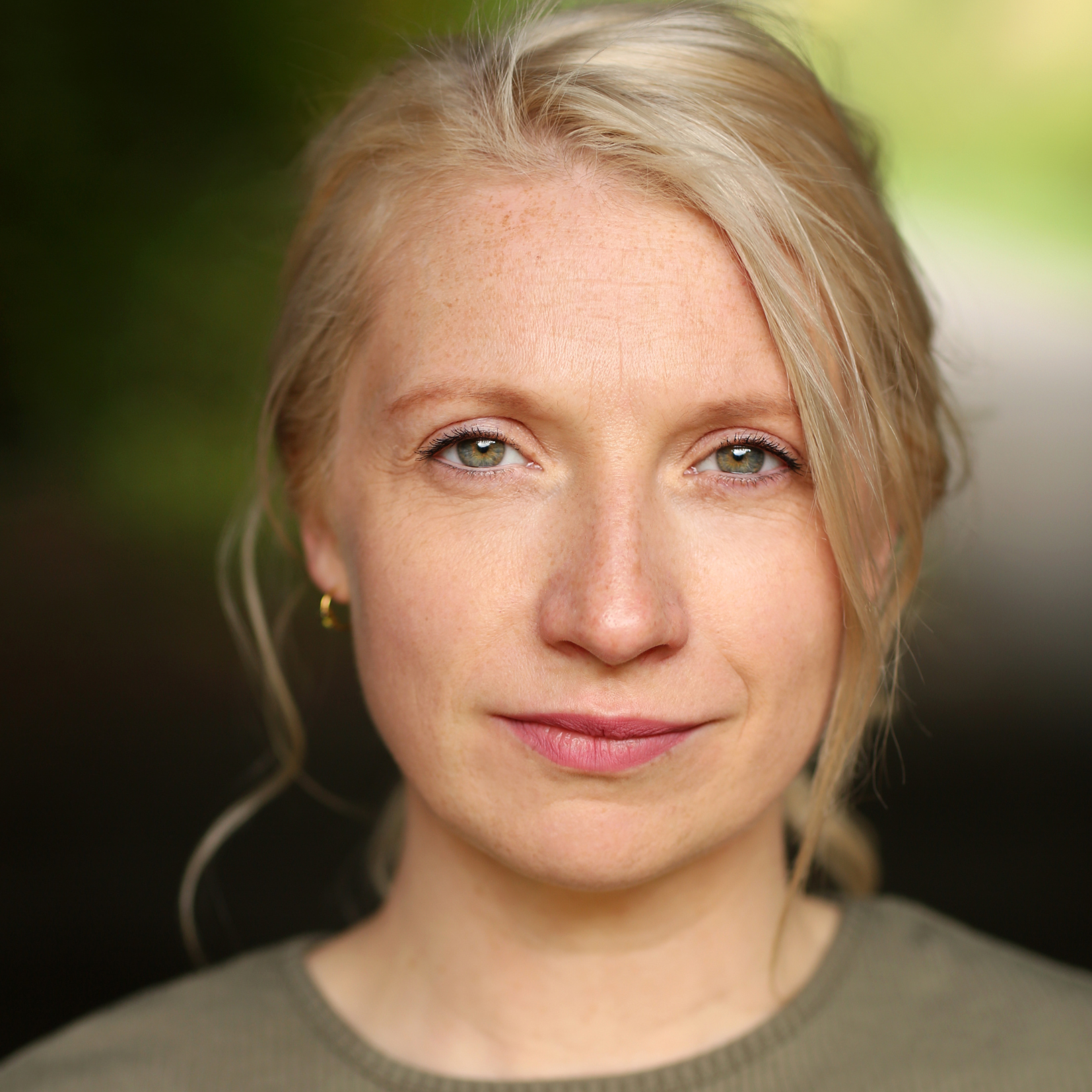 Close-up of a woman with blonde hair and green eyes, smiling softly, wearing a gray sweater.