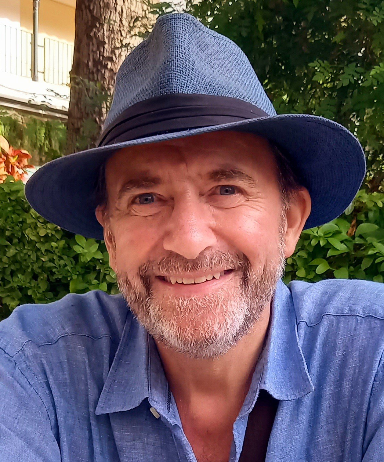 A smiling man with a beard and blue eyes wearing a blue shirt and a large blue hat, outdoors surrounded by green foliage and a tree trunk in the background.
