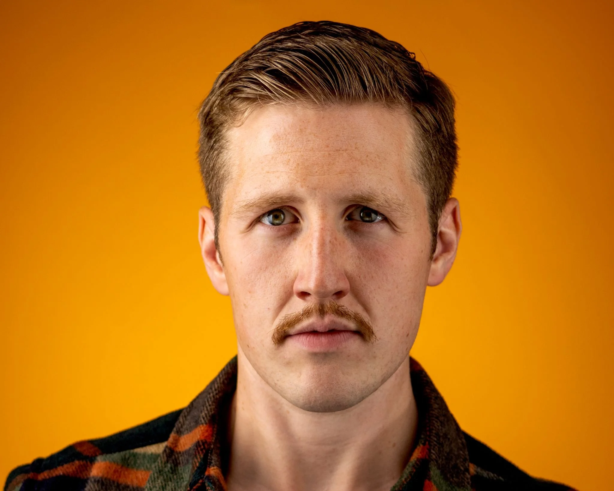 Close-up of a young man with brown hair, green eyes, and facial hair, wearing a patterned jacket, against an orange background.