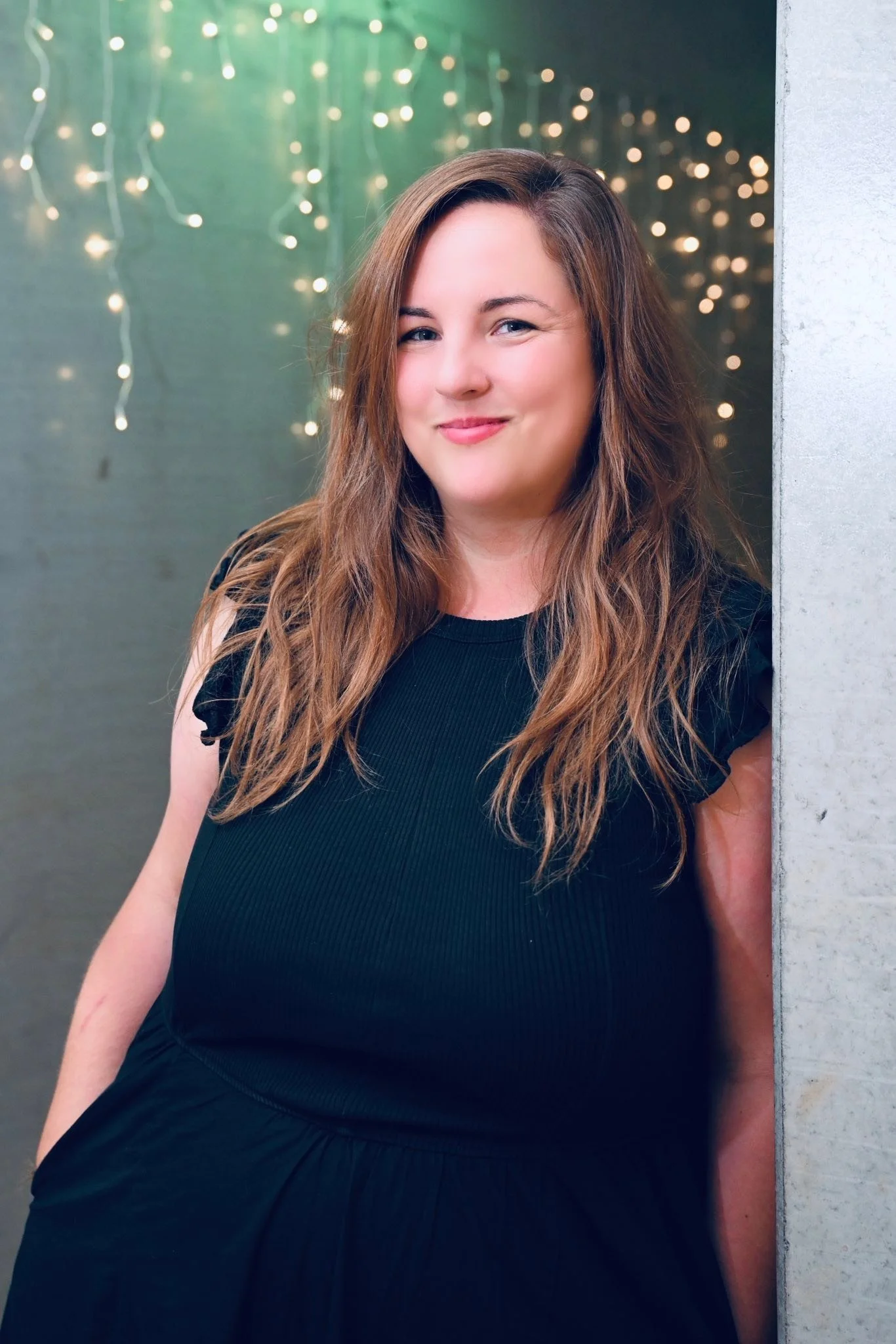 Woman with long brown hair smiling, wearing a black top, standing against a background of blurry string lights and a concrete wall.