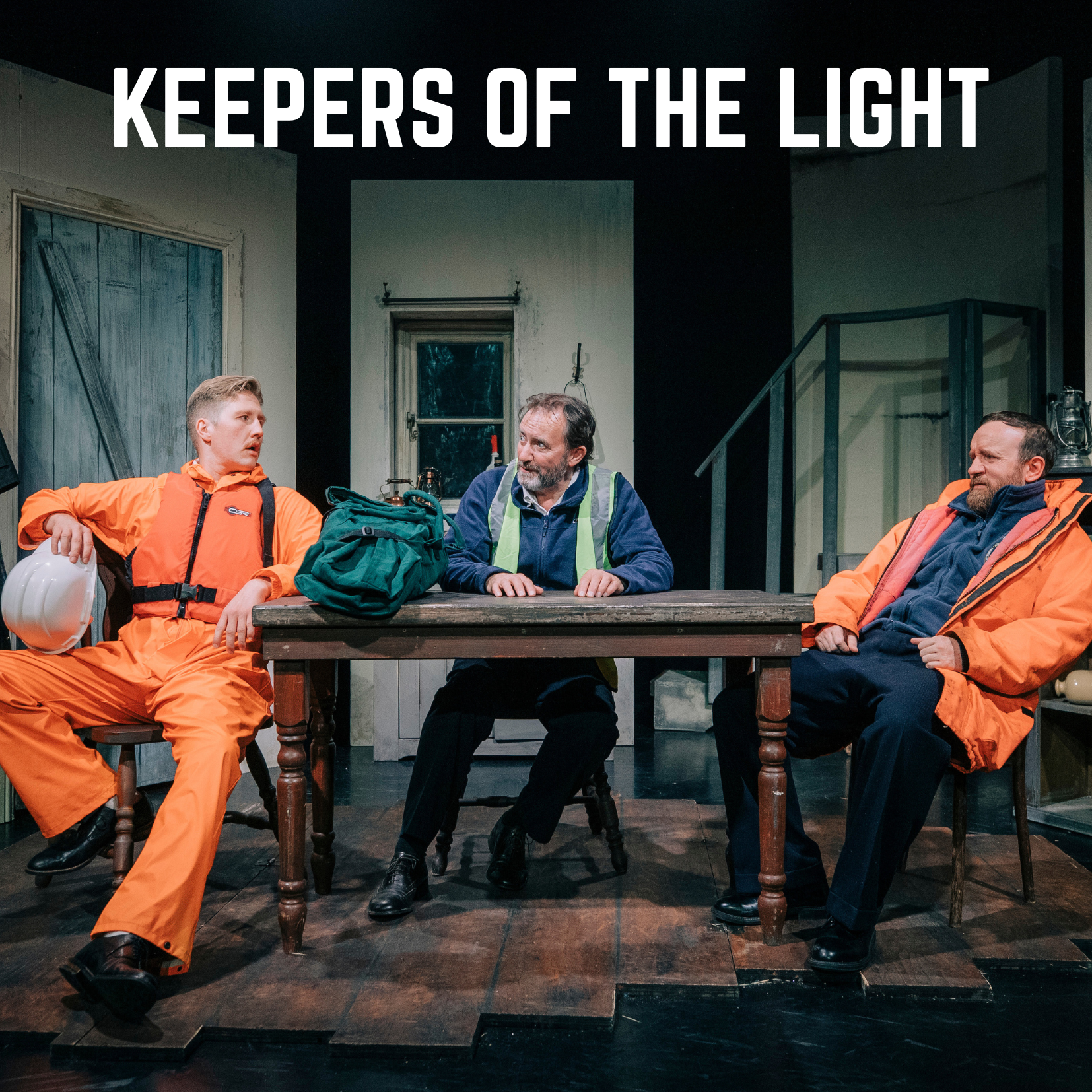 Three men sitting at a wooden table during a play or theatrical performance. The man on the left is wearing an orange jumpsuit and a white helmet, looking to the side. The man in the middle has a beard, wearing a blue vest with reflective strips, and