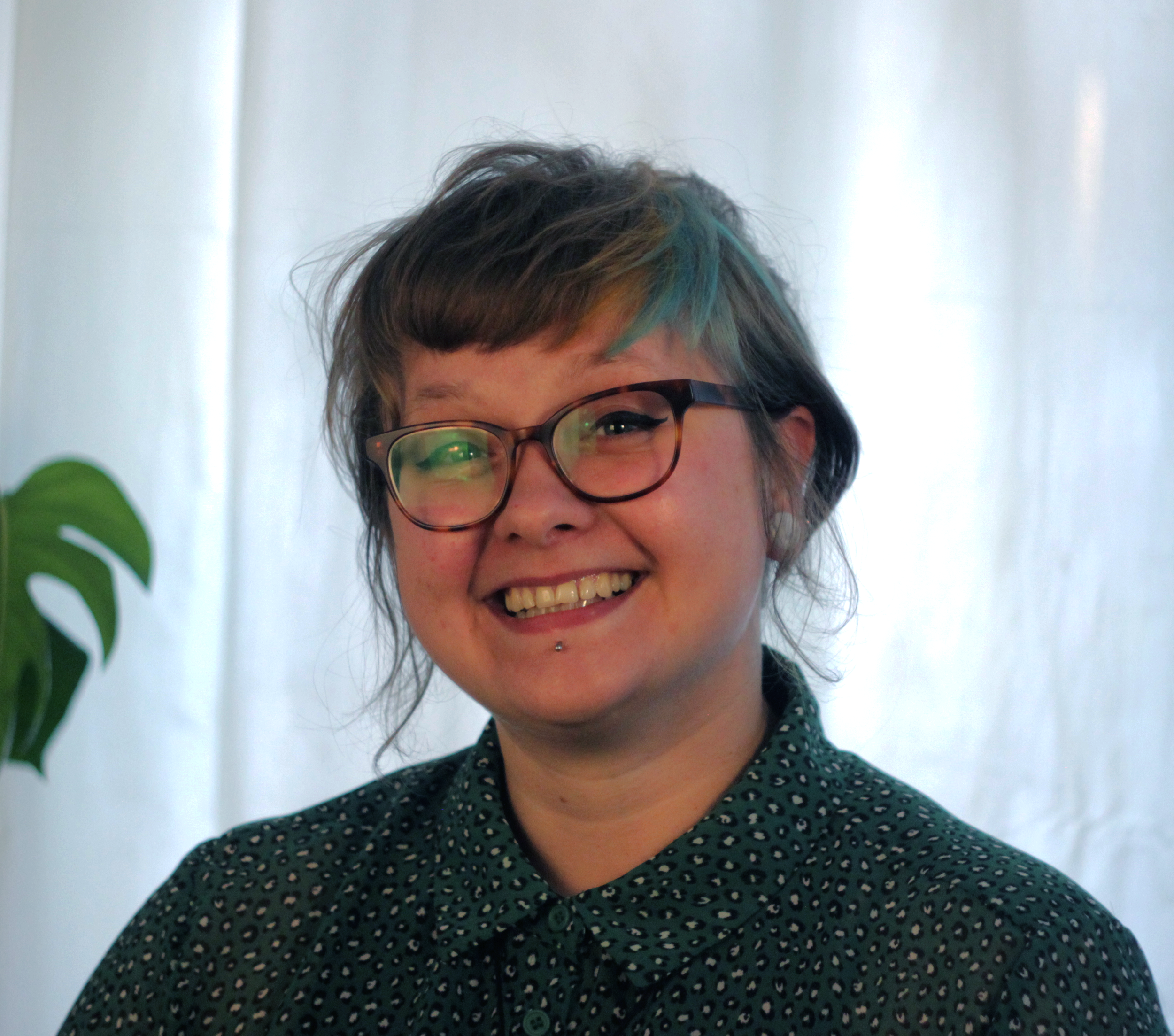 Close-up of a woman with short, curly hair with colorful streaks, wearing glasses, smiling, and wearing a dark green dress with a small red and white pattern, against a light background with a leafy plant visible on the left side.