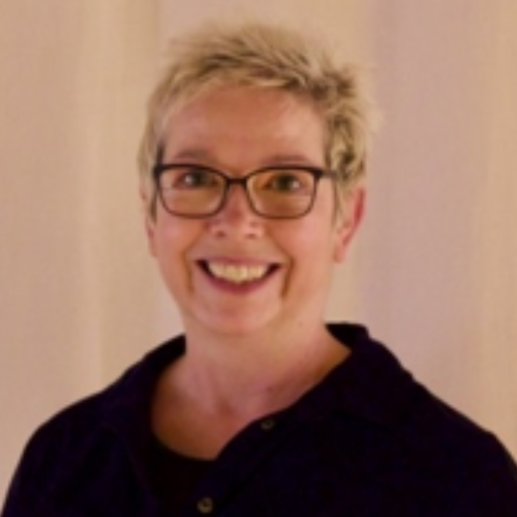 A woman with short blonde hair, glasses, and a black top smiling in front of a beige wall.