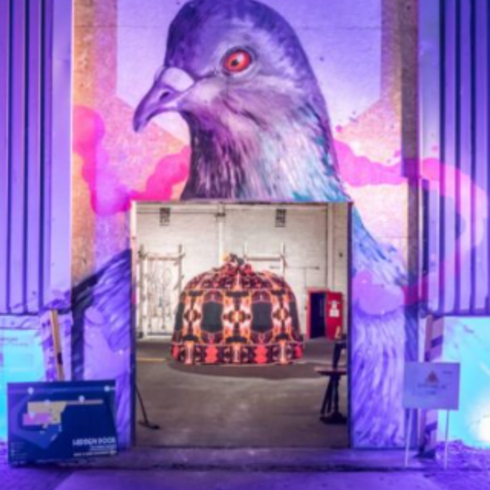 Interior of an art installation featuring a large mural of a bird of prey with red eyes, overlooking a bed with a black, orange, and red patterned blanket. The mural has a purple background with pink clouds, and the room is decorated with purple and blue lighting.