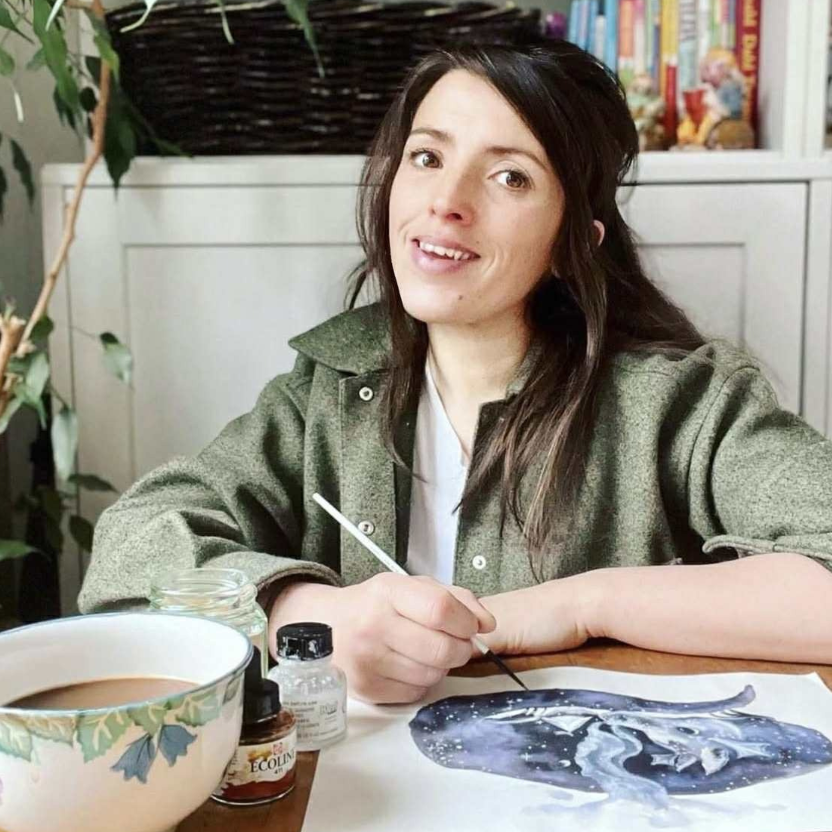 A woman with long brown hair and light skin is painting on a canvas with a paintbrush. She is sitting at a table with assorted art supplies, including a bowl, paint bottles, and a jar labeled 'Ecover'. There is a white cabinet with a basket and colorful books behind her, and a green leafy plant on her left.