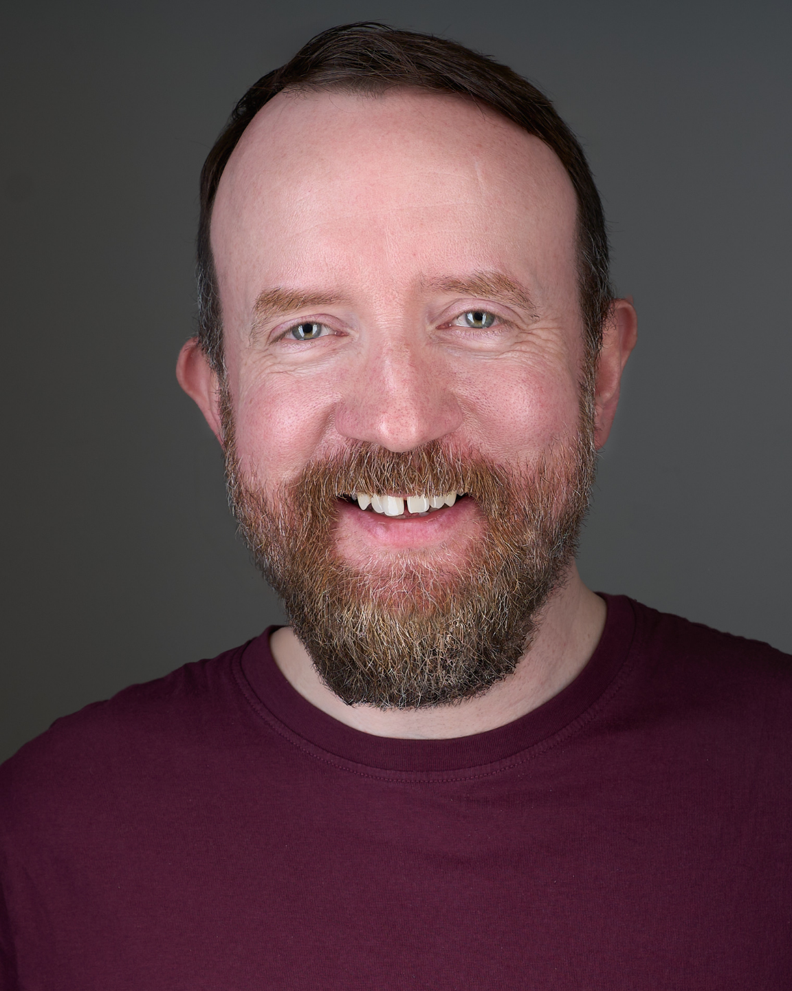 Portrait of a bearded man with light skin, short brown hair, wearing a maroon shirt, smiling against a gray background.