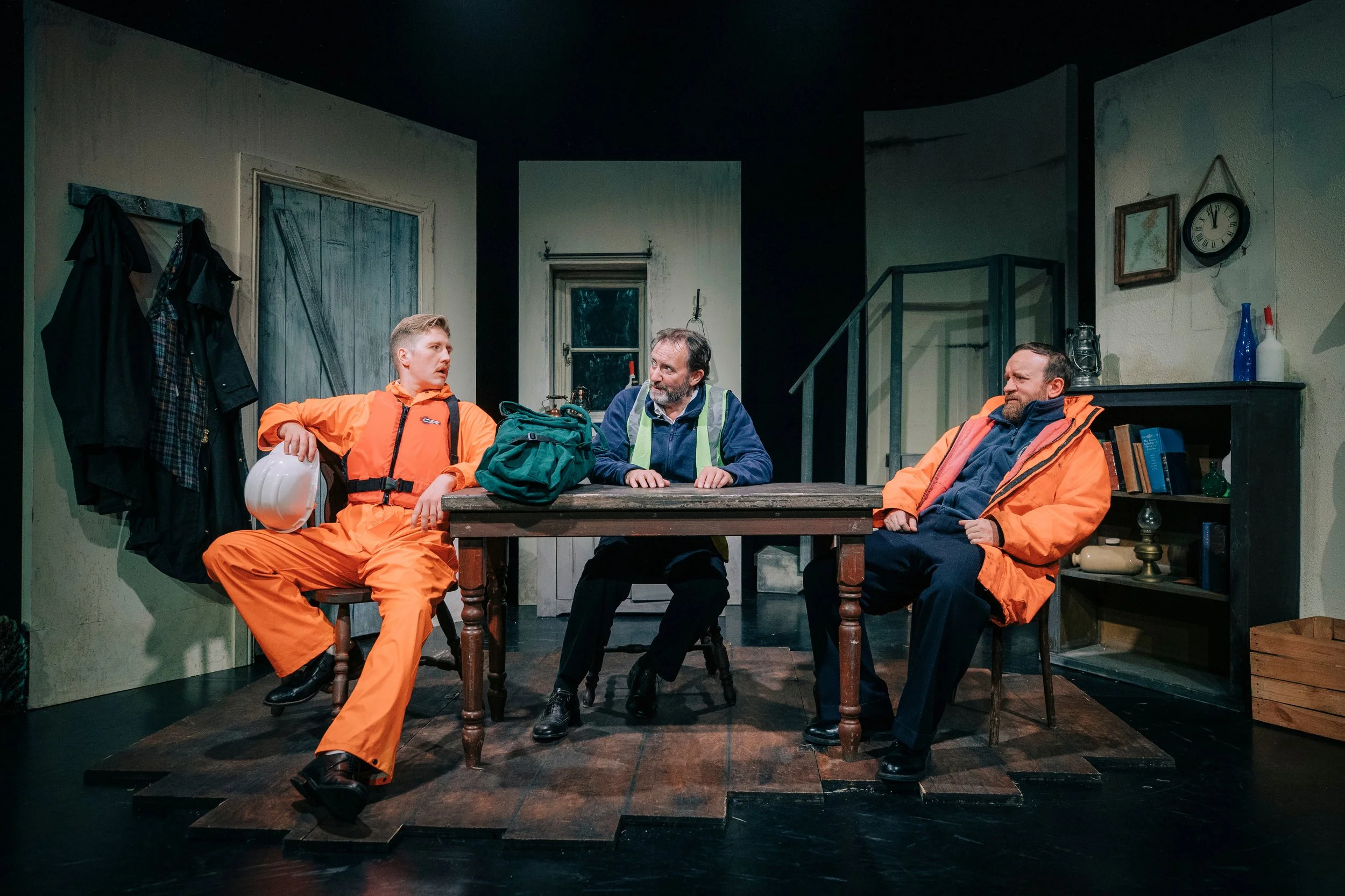 A scene from a theatrical play showing four actors in a room with worn walls and simple furniture. Two actors, dressed in bright orange outfits with safety vests, sit at a wooden table. One holds a helmet, and the other is leaning back with hands on 