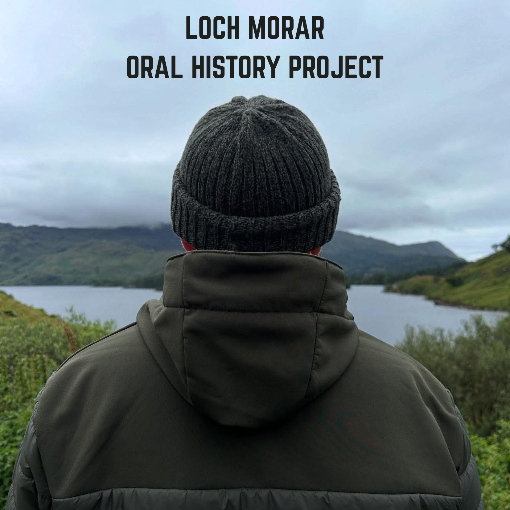 Person wearing a gray knit beanie and dark gray jacket, facing away in a scenic outdoor setting with loch, hills, and cloudy sky in the background.