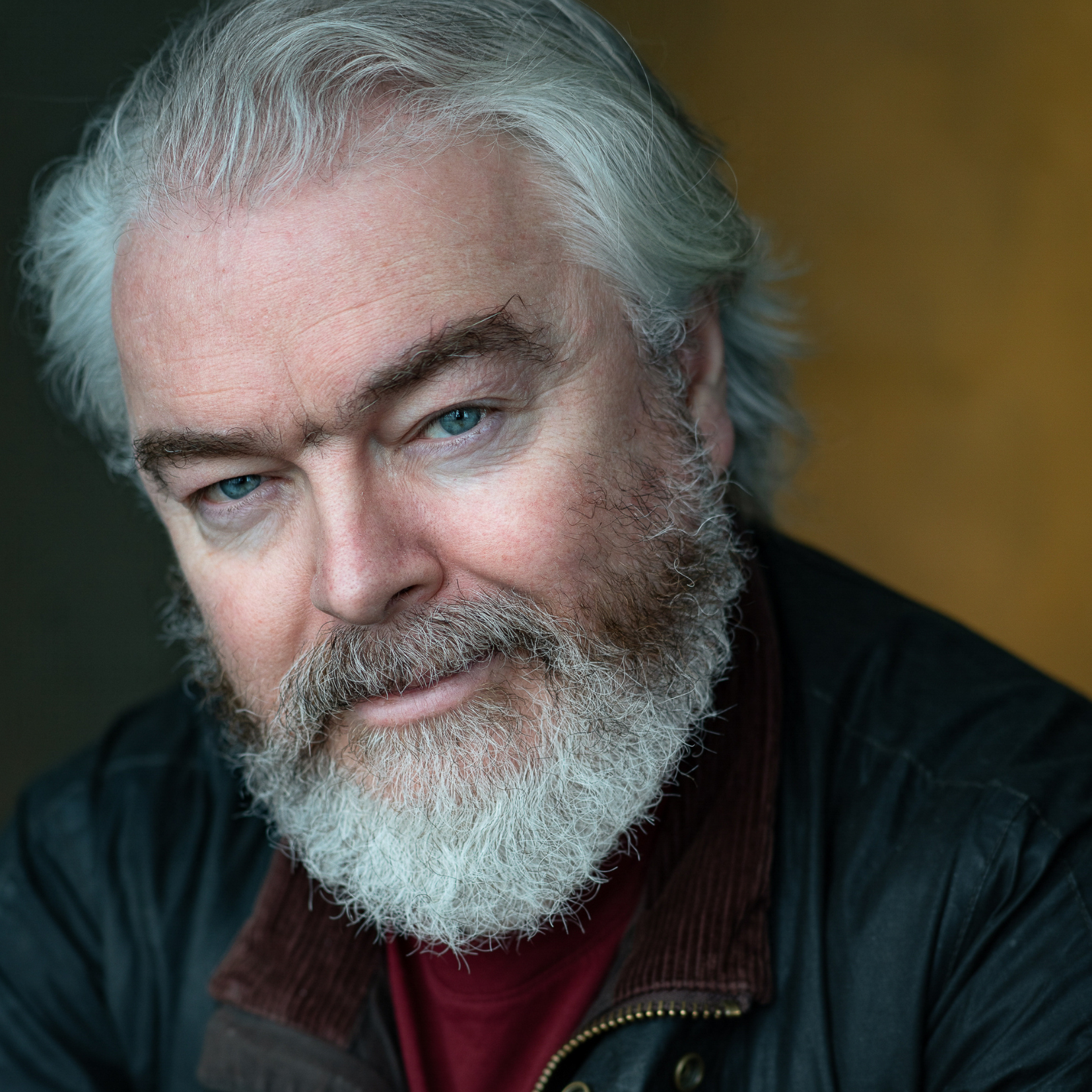 Close-up of an older man with gray hair, blue eyes, and a white beard, wearing a dark jacket with a gold zipper and a maroon scarf.