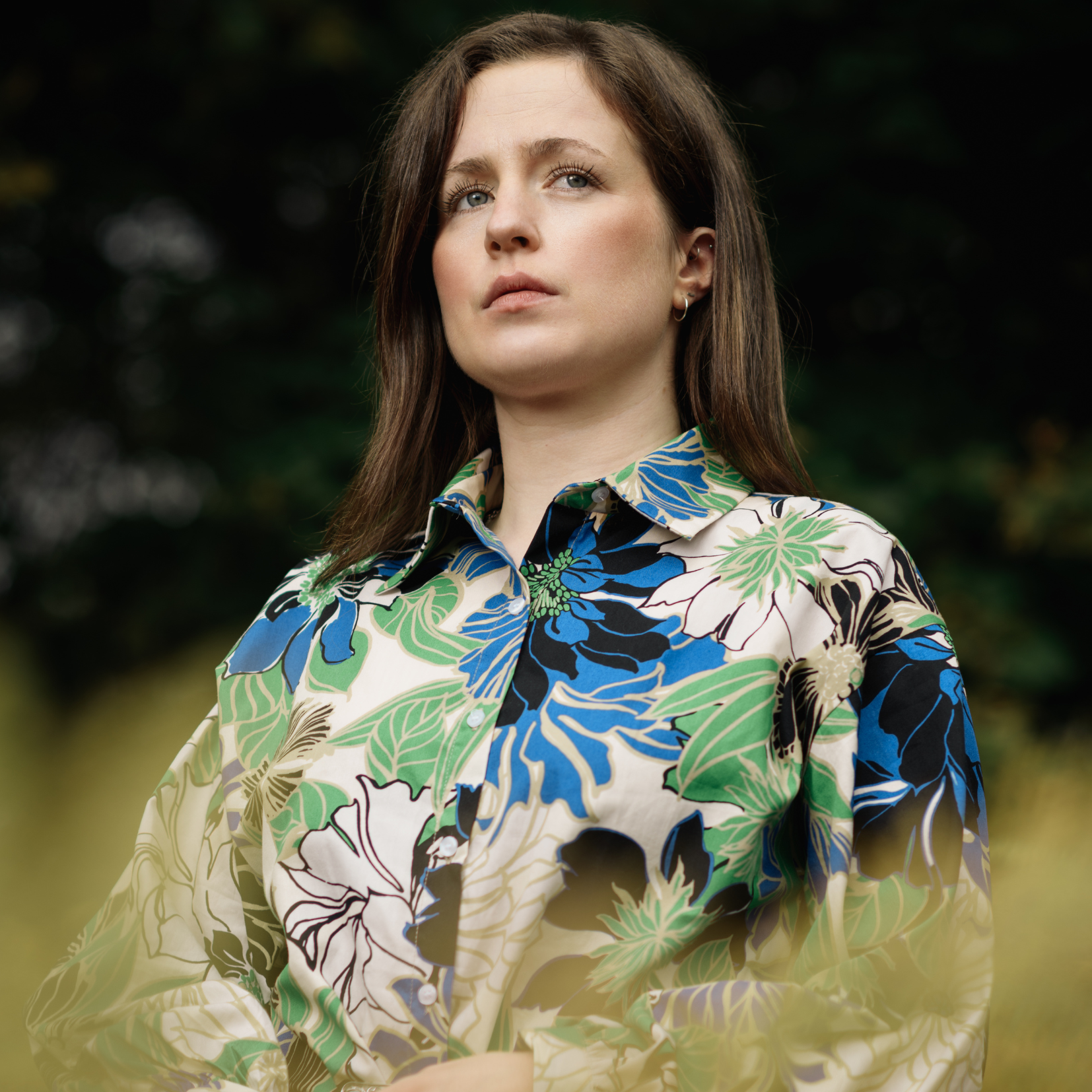 A woman with brown hair and blue eyes wearing a colorful floral patterned shirt outdoors with dark blurred foliage in the background.