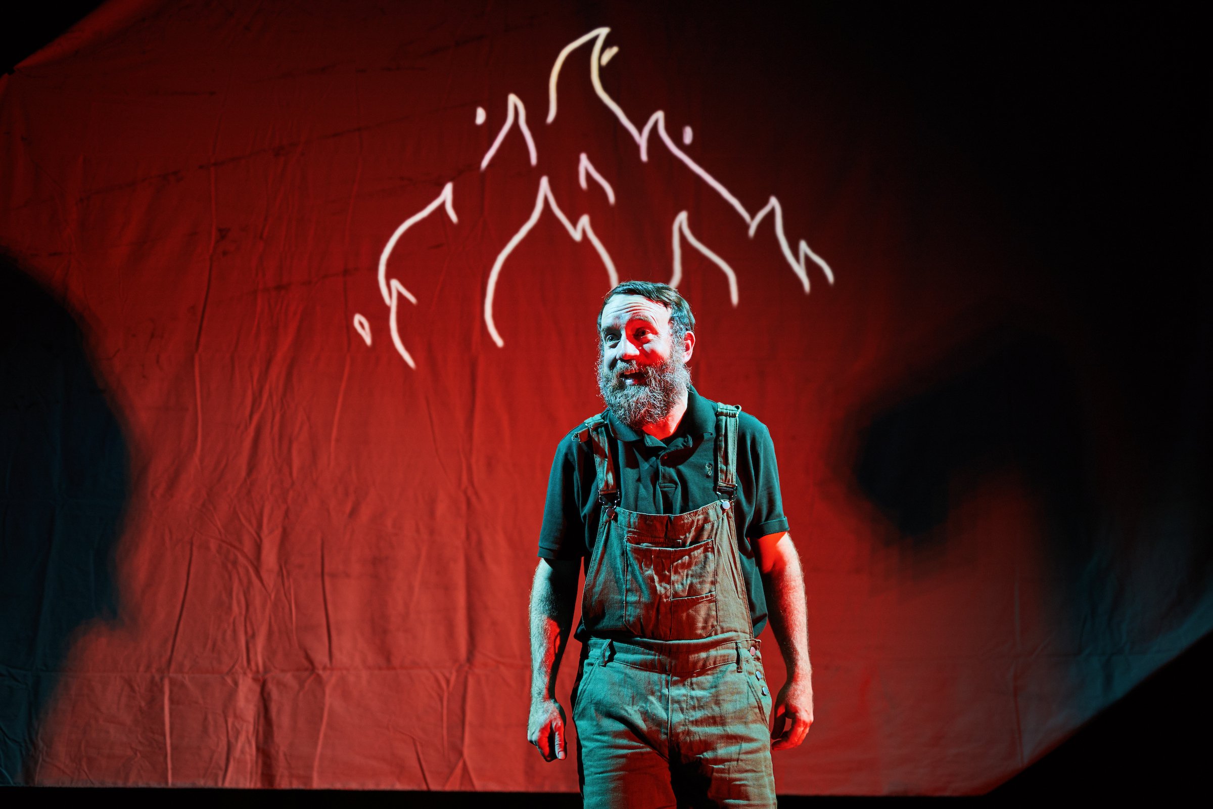A man with a beard wearing overalls standing in front of a red background with a white neon outline of a mountain above him.