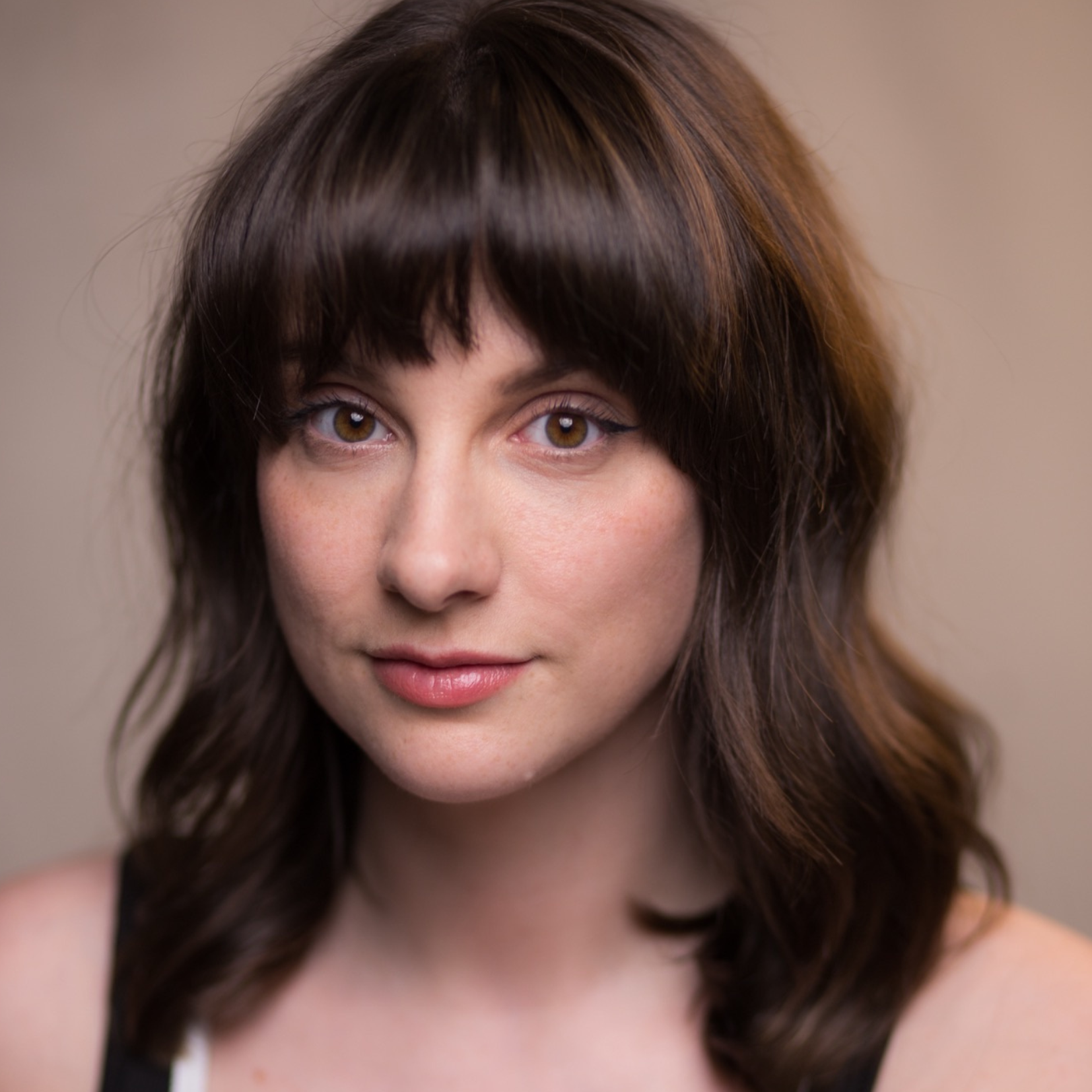 Close-up portrait of a woman with hazel eyes, brown hair with subtle highlights, and a gentle smile, against a plain beige background.