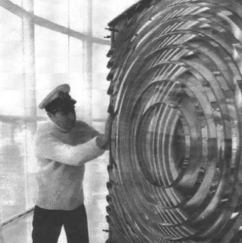 A man dressed as a sailor stands next to a large, spiral-shaped metallic sculpture indoors. He appears to be touching or adjusting the sculpture.