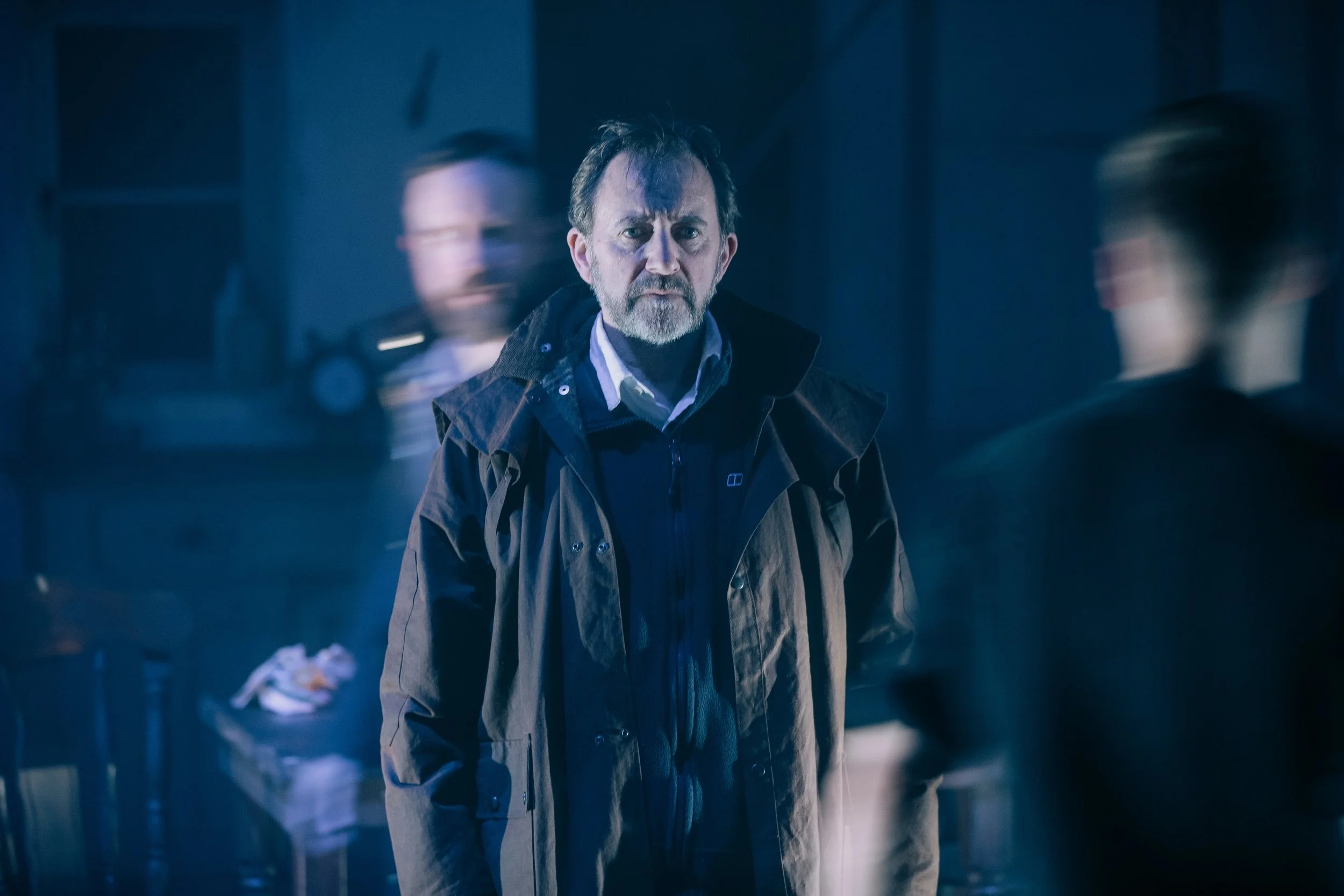 A man with a beard and short hair wearing a dark jacket and light-colored shirt, standing in a dimly lit environment with blurred human figures in the background.