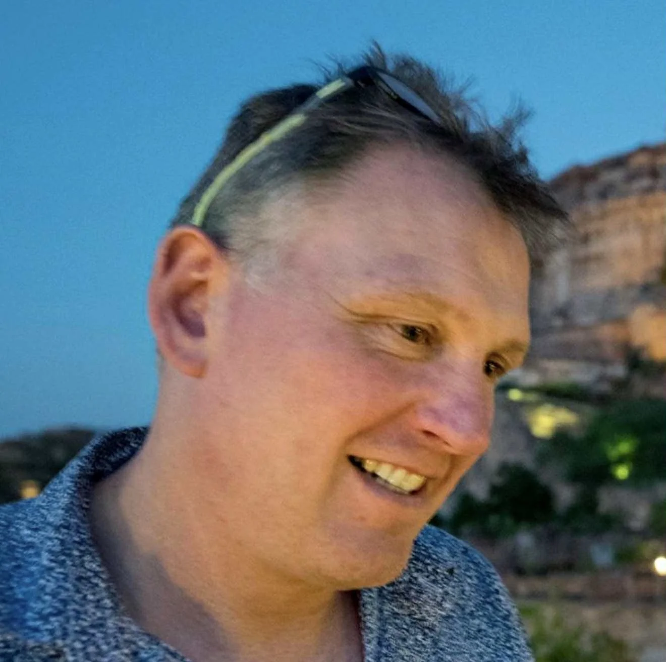 A smiling person with glasses on their head, outdoors at dusk with mountains and trees in the background.