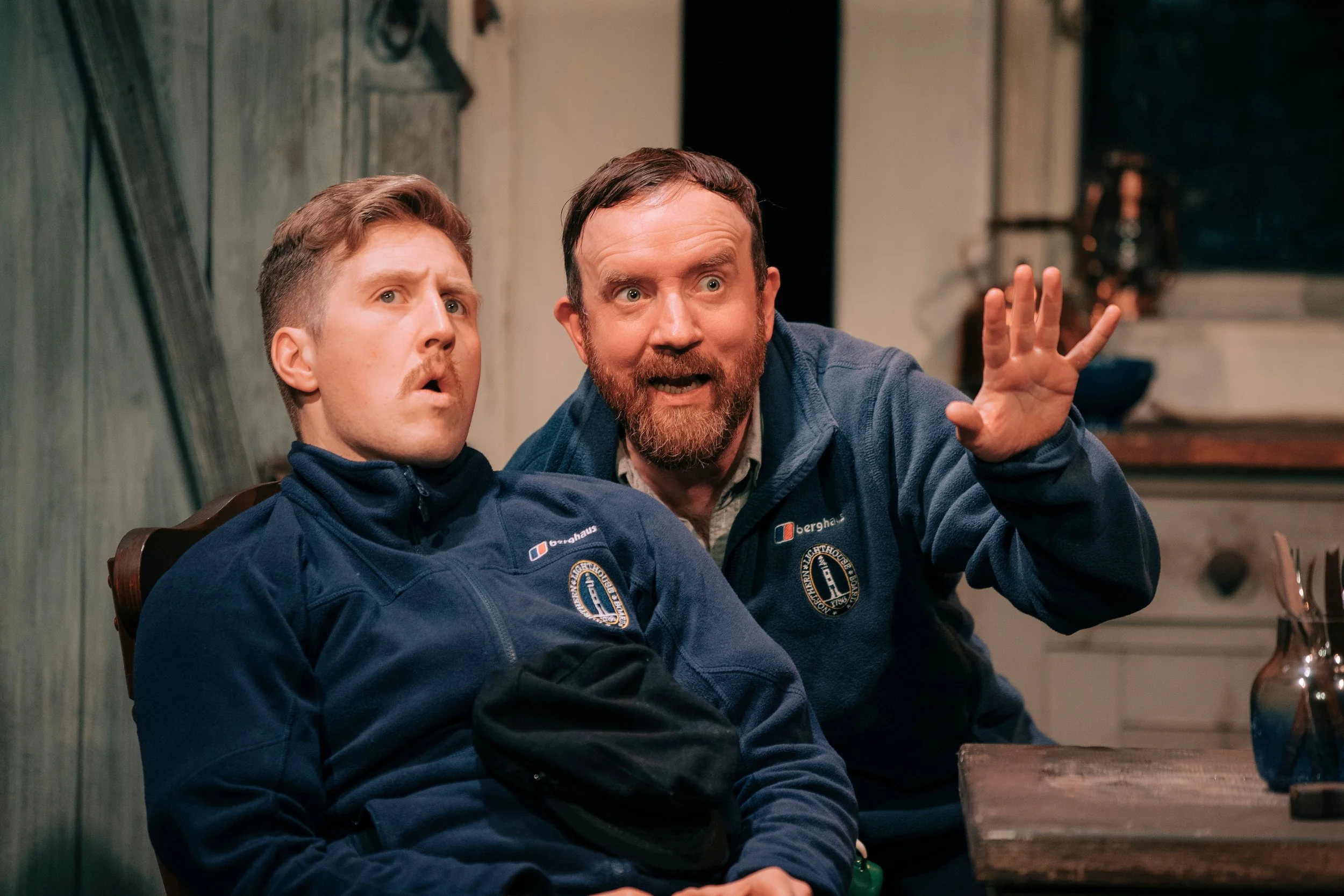 Two men wearing dark blue jackets with patches on the sleeves sitting at a wooden table in a rustic room. One man has a beard and is gesturing with his hand, both appear to be engaged in a conversation.