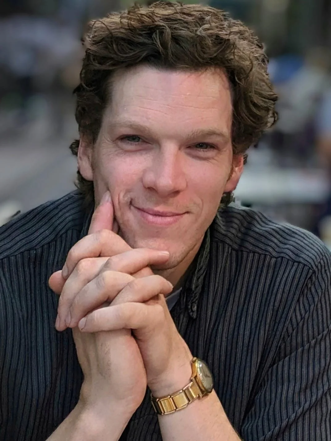 A man with curly brown hair, wearing a watch and a dark striped shirt, sitting with his hands clasped near his face and smiling.