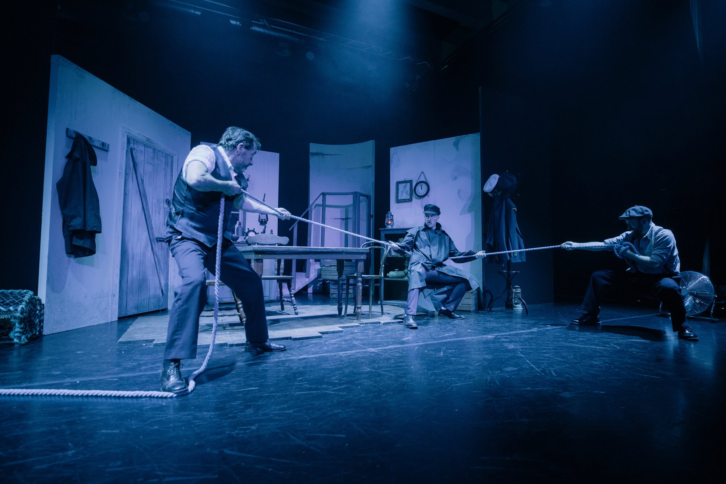 Three men on stage pulling on a rope during a theatrical performance. The stage has simple decor including a clock, picture frames, and a door. The scene is lit with blue lighting.