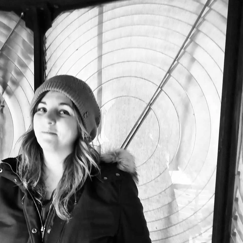 A young woman wearing a beanie and a winter coat stands in front of an archery target with an arrow stuck in the bullseye.