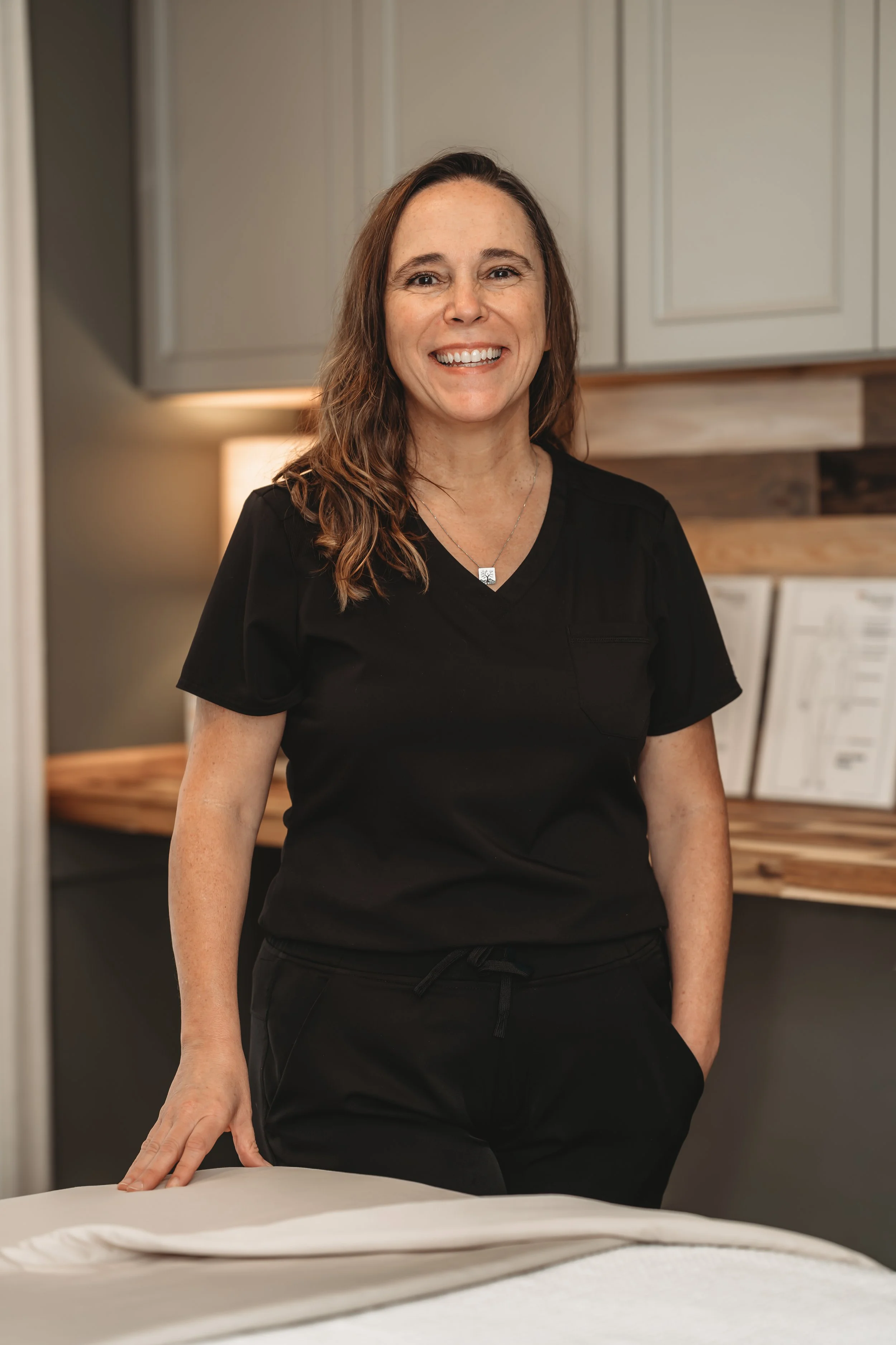 Owner Serena standing behind a massage table, smiling, with cabinets in the background.