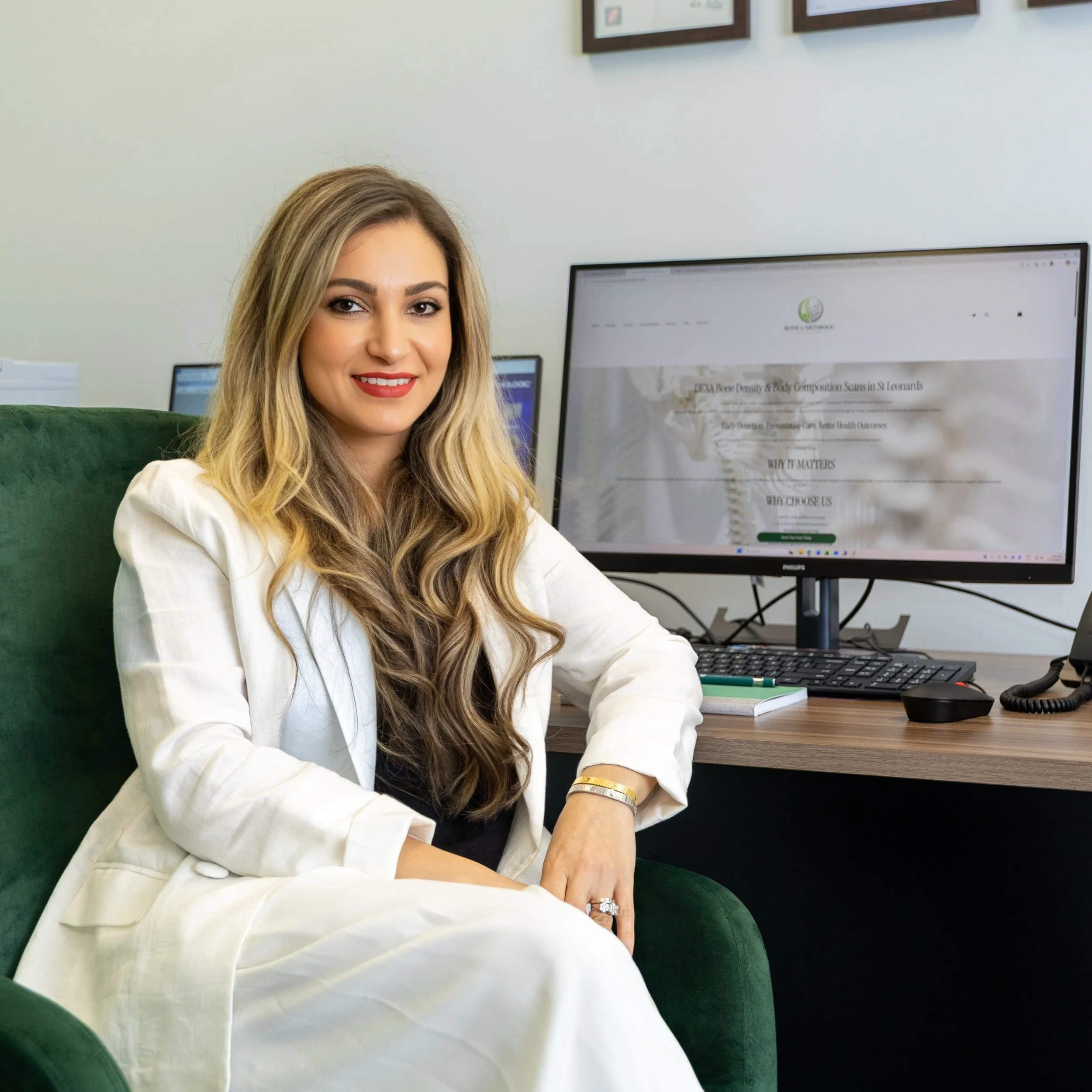 A woman with long blonde hair and red lipstick sitting in a green chair in front of a computer monitor in an office setting.