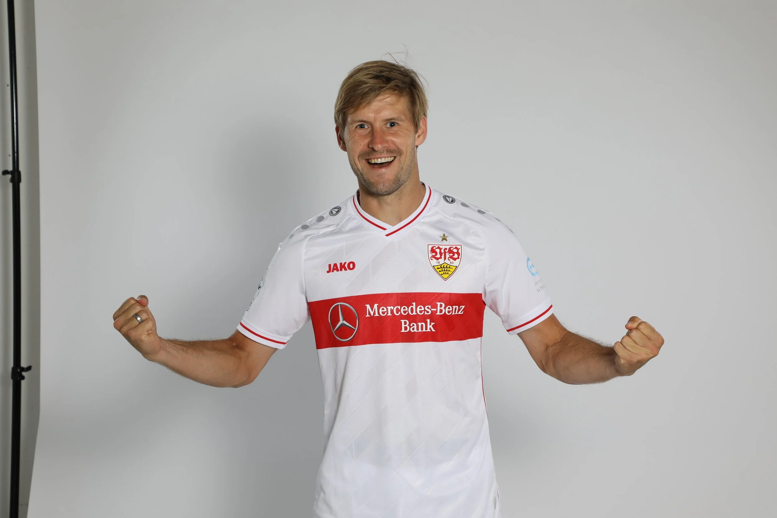 Ein männlicher Mensch trägt ein Weißes Fussballtrikot mit roten und schwarzen Akzenten, das die Logos von Mercedes-Benz Bank, JAKO und VfB Stuttgart zeigt, und lacht freundlich, während er seine Arme anspannt.