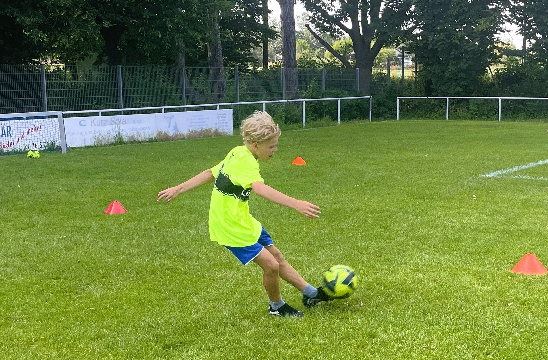 Ein Jugendlicher beim Fußballtraining auf einem grünen Rasen, umgeben von einem weißen Zaun und Bäumen im Hintergrund, mit orangefarbenen Hütchen auf dem Boden.