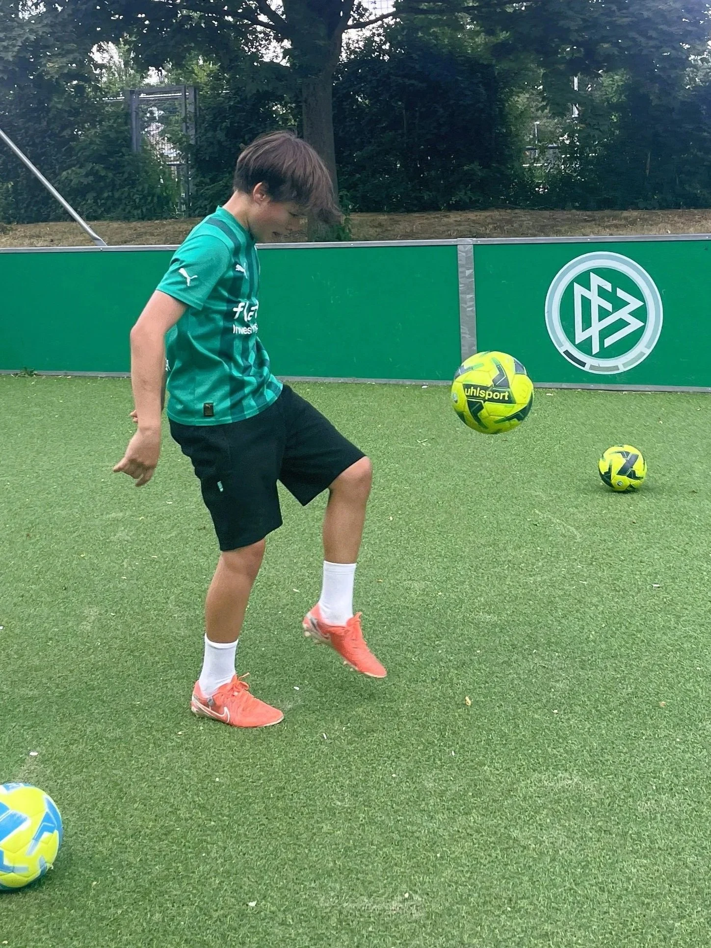 Ein Junge beim Fußballspielen auf einem Sportplatz, trägt ein grünes Trikot, schwarze Shorts, weiße Socken und orange Fußballschuhe, steht vor einem Ball, im Hintergrund ein grüner Zaun mit einem Logo und Bäumen.
